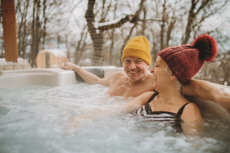 Senior couple in kintted cap enjoying together outdoor bathtub at their terrace during cold winter day.