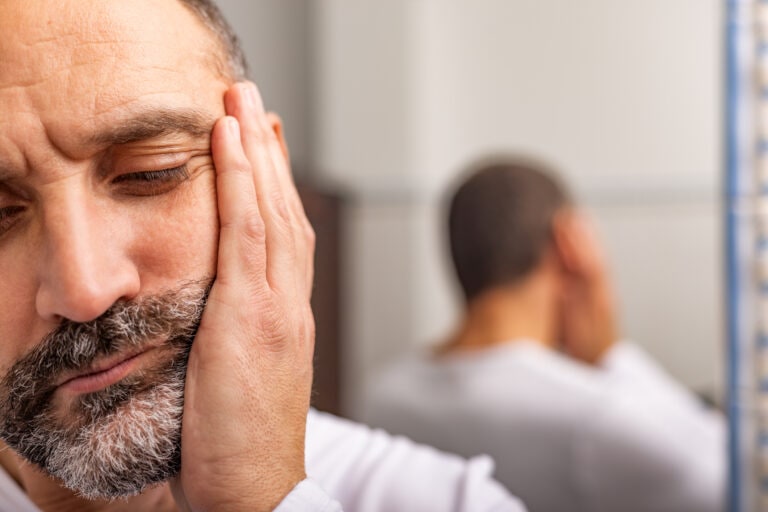 Close-up of adult bearded man's face with toothache. In the background, his reflection in the bathroom mirror.
