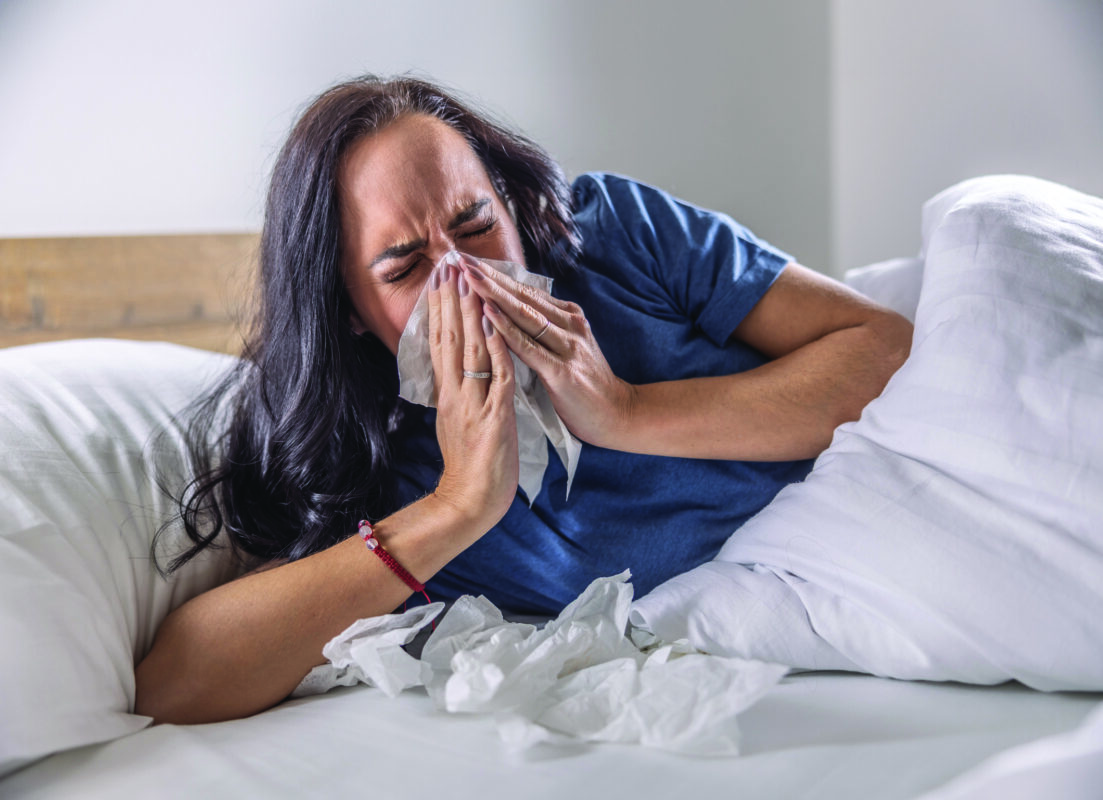 A sick woman sneezes into a napkin, lies in bed, has the flu or a cold.