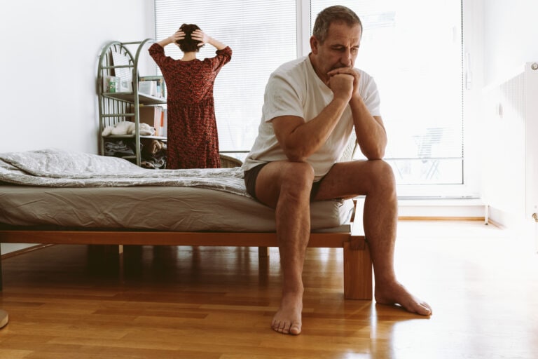 middle-aged man sits at end of bed and holds head after an argument with wife standing at window, view from back.