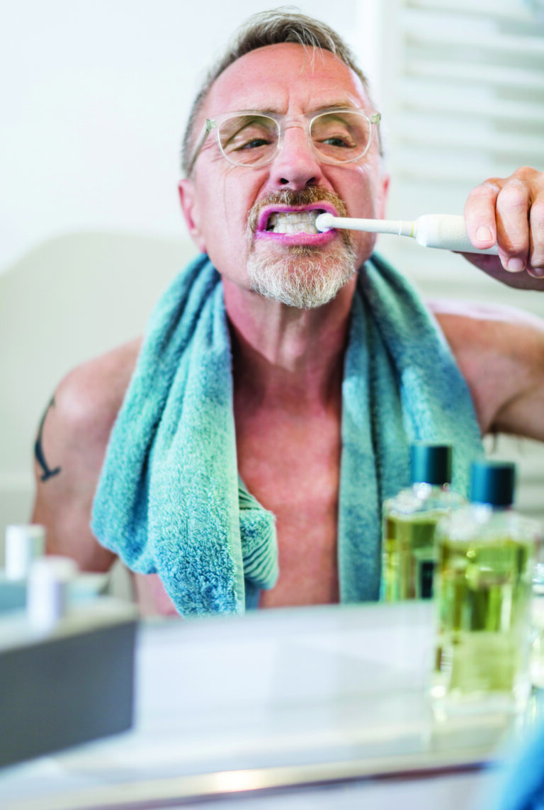Senior handsome and shirtless man with towel around his neck in his 60s is brushing his teeth while looking in mirror.