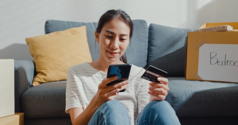 Young Asian woman moving house sit on floor holding smartphone and banking credit card after unpacking.